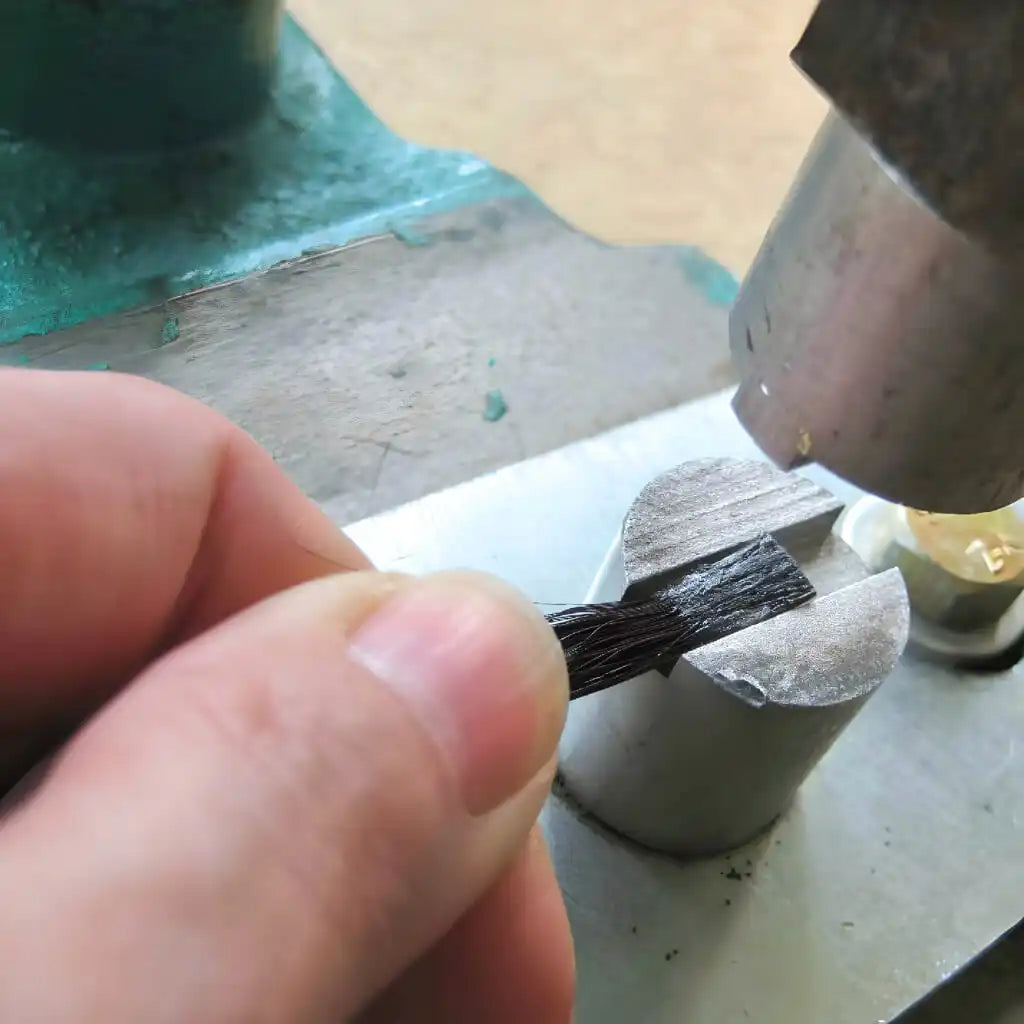 Eine Hand hält ein Bündel schwarzer Fasern, die über einer Metallschraube positioniert sind, während ein Metallobjekt Druck ausübt.